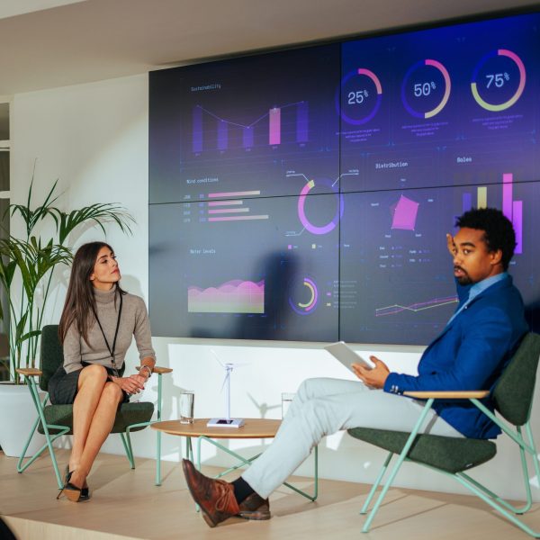 A specialist is presenting a lecture to a full auditorium of businesspeople in a conference hall with the use of a digital tablet and charts on a led display.